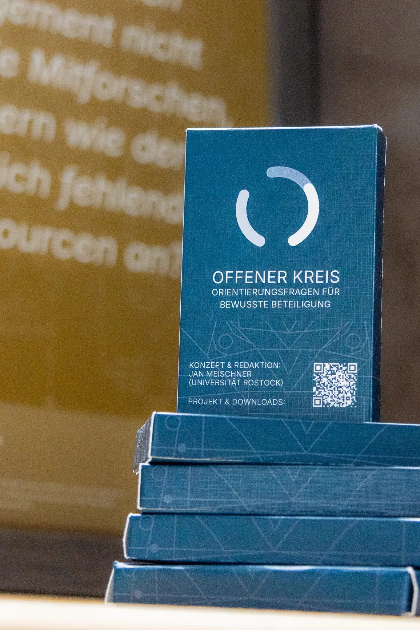 Several printed Offener Kreis card sets stacked on a table, with one standing upright in front of a blurred project poster.